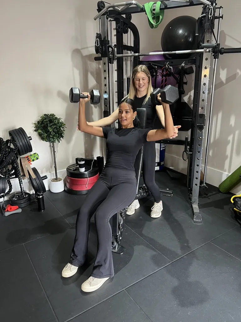Ella supporting a client through a seated dumbbell press in the studio gym