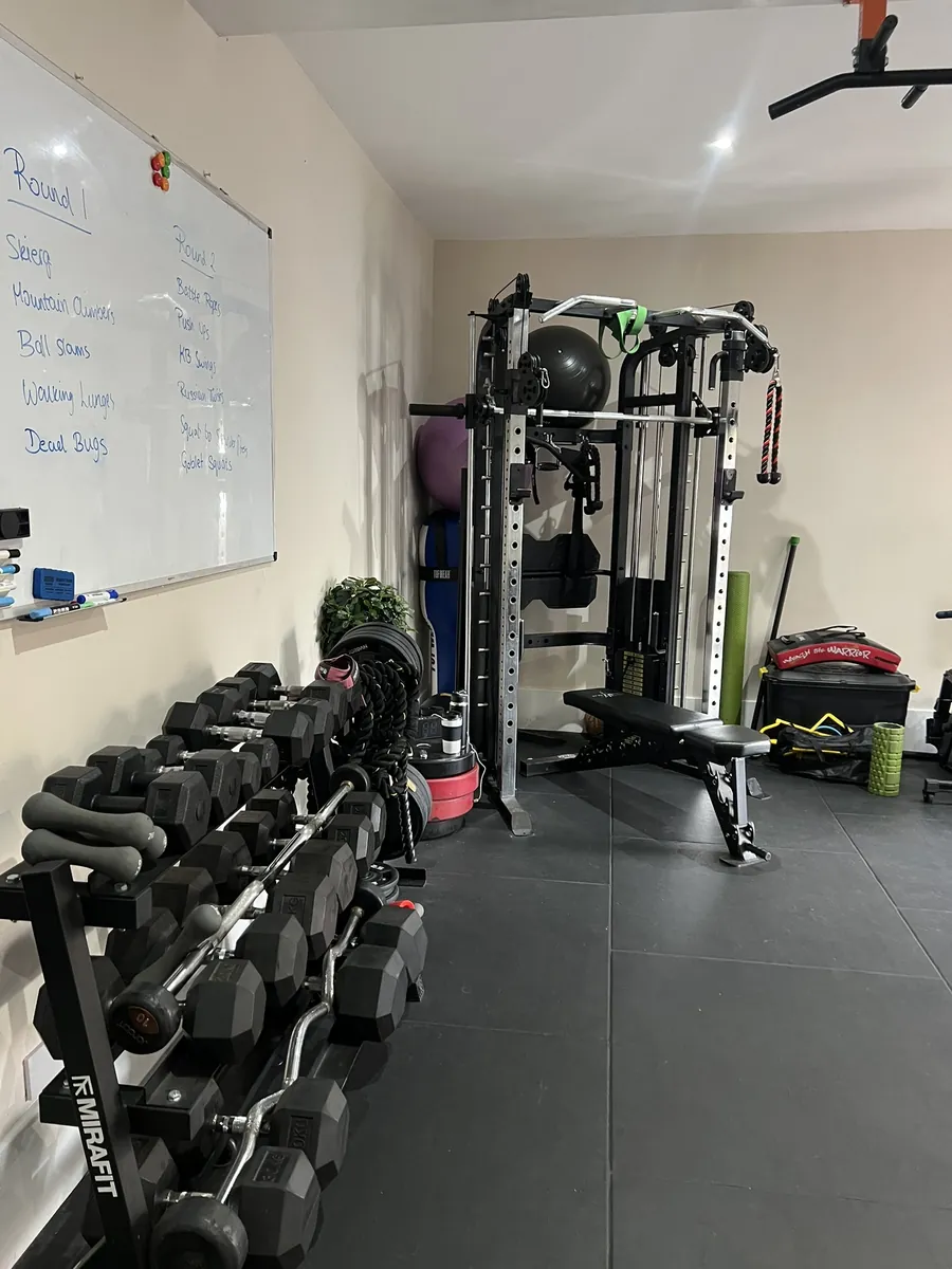 The studio floor, with a dumbbell rack, cable machine, bench and the class whiteboard